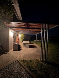 Famille sous pergola bois éclairée de nuit, terrasse moderne avec mobilier en résine tressée grise.