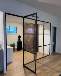 Verrière d'atelier noire en métal ouvrant sur une pièce avec parquet clair et armoire ancienne.